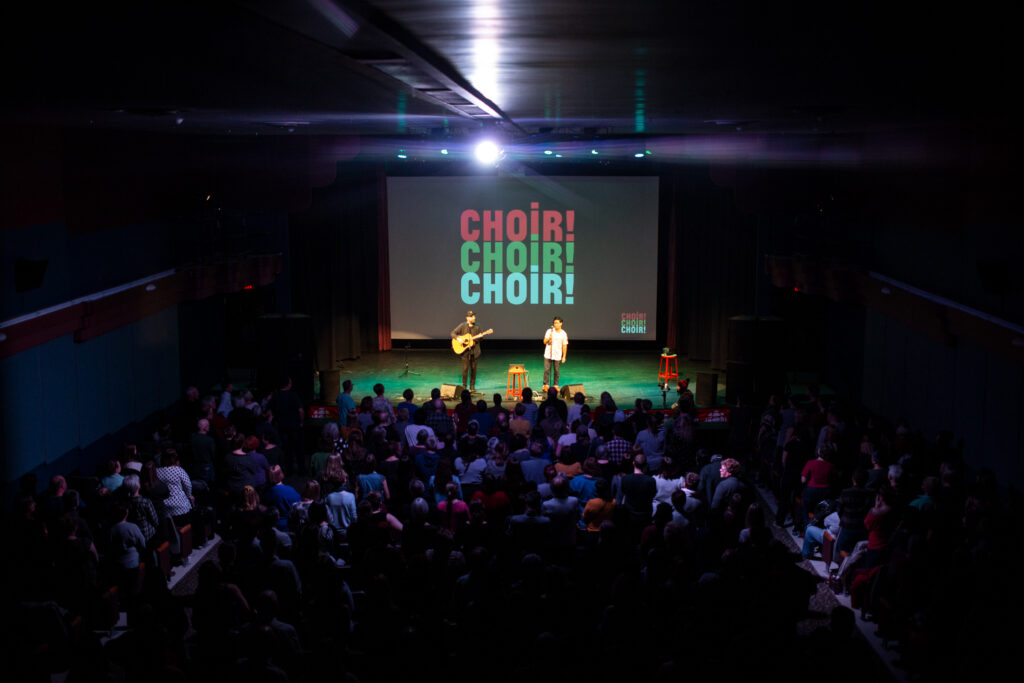 This coloured photo shows a full theatre of people focused on two musicians on stage in front of a 'Choir!Choir!Choir!' on screen.