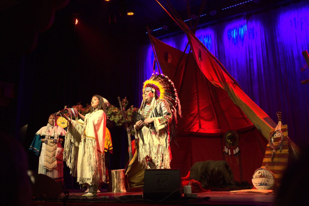 This coloured photo shows three performers in regalia in front of a tipi and other historical artifacts.
