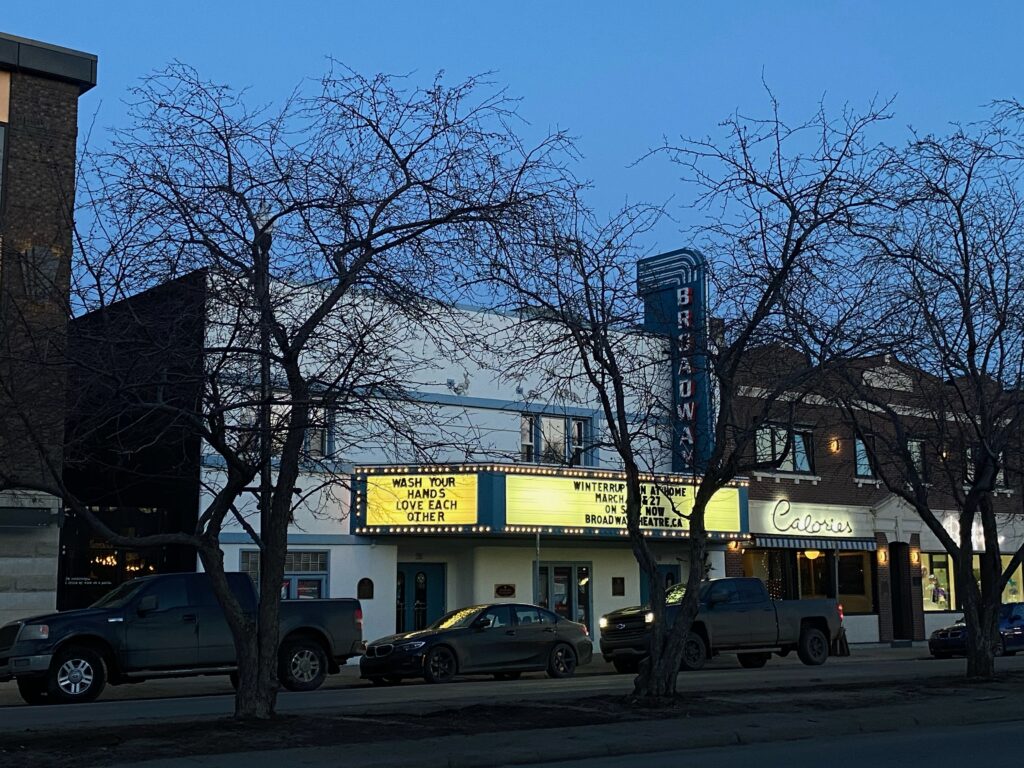 A coloured photo shows the Broadway Theatre at dusk with the marquees lit up focusing on the sign 'Wash your hands, love each other,' There are three threes in front of the Theatre as well as a few cars.