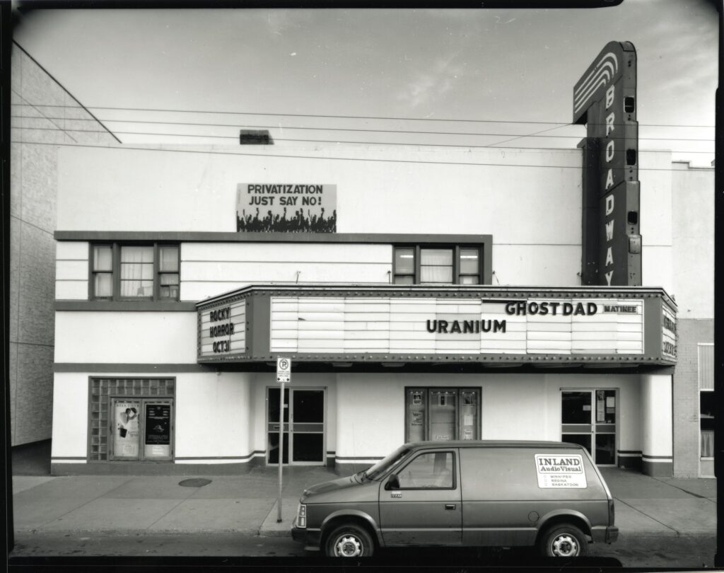Black and white photo with the white-walled Broadway Theatre exterior centred. A mural on the upper half of the building as well as marquees displaying upcoming films. A van is parked in front with an'Inland Audio Video' sticker on the back.